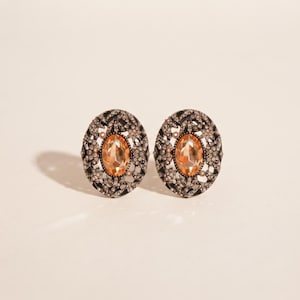 May include: A pair of oval-shaped earrings featuring a central peach-colored gemstone surrounded by a dark, intricate filigree metal frame encrusted with small, clear sparkling crystals arranged in a detailed, textured pattern.