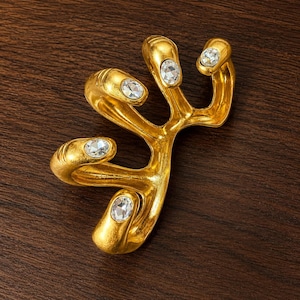 May include: A gold-colored, abstract brooch with a branching design. The brooch features five clear, oval-shaped gemstones. The jewelry piece is set against a dark brown wooden surface.