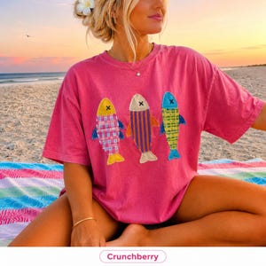 May include: A pink short-sleeved t-shirt featuring three fish appliques on the front. The left fish has a pink and white plaid pattern, the centre fish has vertical purple and orange stripes, and the right fish has a green and yellow plaid pattern.