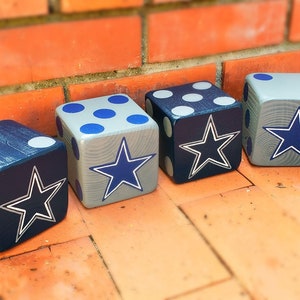 May include: Four wooden dice, painted in navy blue and gray, with white and blue dots and a white-outlined blue star. The dice are arranged on a terracotta tile surface, leaning against a brick wall.