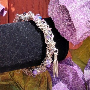 Puede incluir: Una pulsera de alambre de metal plateado, con pequeñas cuentas moradas y transparentes tejidas en la estructura. Un pequeño dije colgante en forma de pluma de metal plateado está sujeto al extremo de la pulsera.