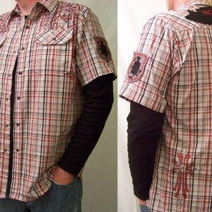 May include: A short-sleeved button-down shirt featuring a red, white, and black plaid pattern. The shirt includes two chest pockets, red embroidery on the shoulders, a patch on the left sleeve, and a large red cross graphic on the lower back.