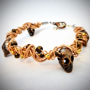 Può includere: Un bracciale realizzato con filo di rame avvolto, caratterizzato da diversi ciondoli a forma di teschio in metallo scuro, intervallati da piccole perline rotonde marrone scuro e minuscoli accenti dorati, chiuso da un moschettone.