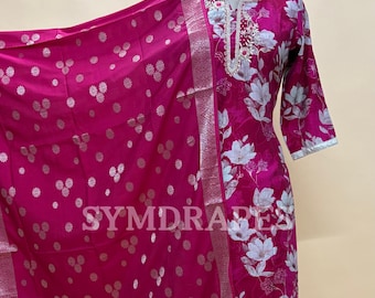 May include: A magenta salwar kameez set with a floral print and a matching dupatta. The outfit features white floral accents and silver trim. The dupatta has a silver paisley border. The text "SYMDRARES" is visible.