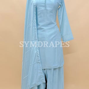 May include: A light blue three-piece outfit, including a long-sleeved top, wide-leg pants, and a matching scarf. The outfit is displayed on a mannequin. The text "SYMDRARES" is visible on the outfit. The outfit is made of a lightweight fabric.