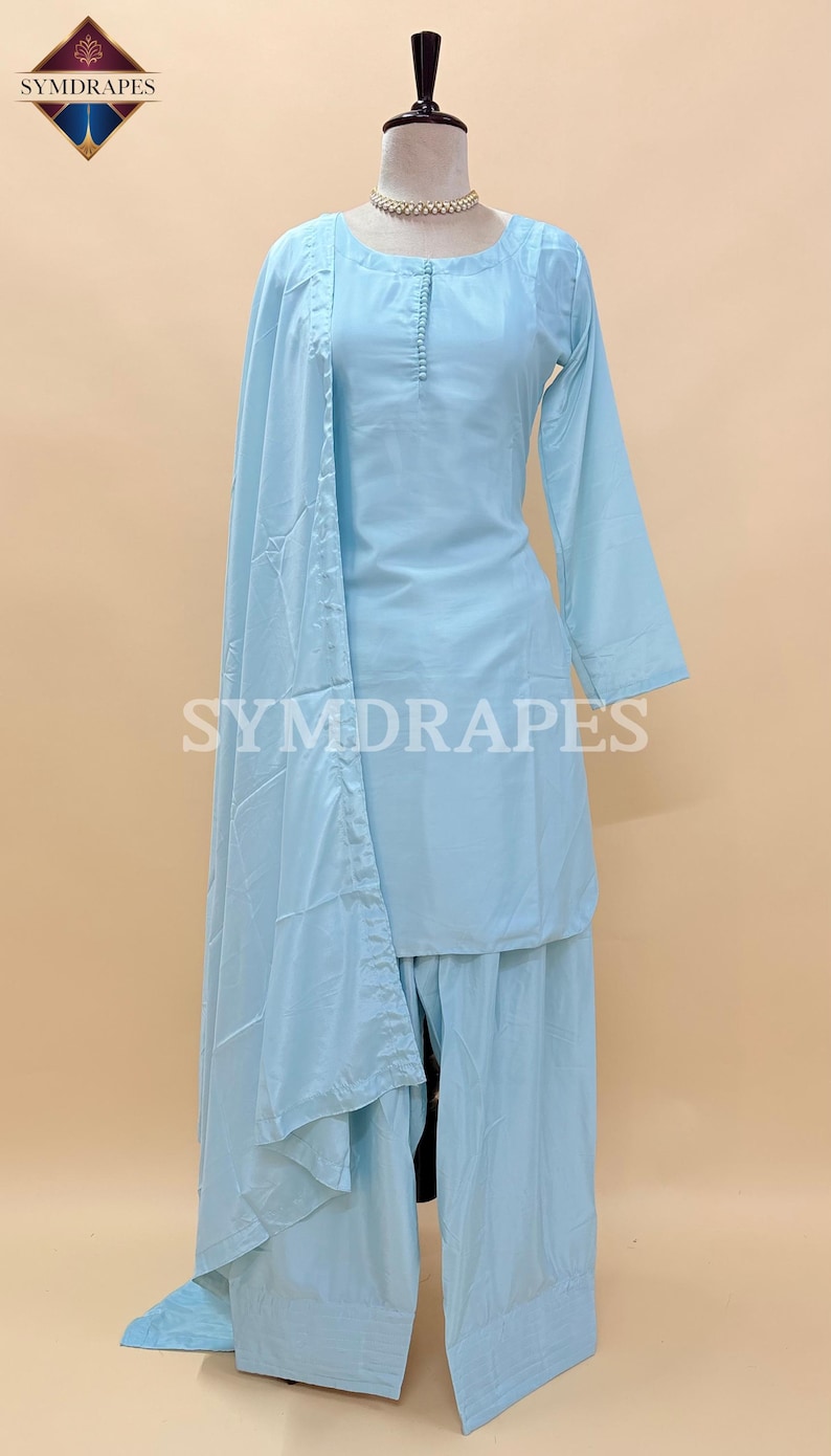 May include: A light blue three-piece outfit, including a long-sleeved top, wide-leg pants, and a matching scarf. The outfit is displayed on a mannequin. The text "SYMDRARES" is visible on the outfit. The outfit is made of a lightweight fabric.