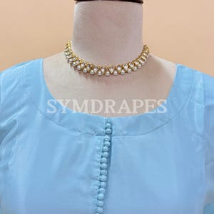 May include: A light blue tunic-style top with a row of small buttons down the front. A gold-colored necklace with white pearl accents is around the neckline. The text "SYMDRAPES" is visible on the top. The top is displayed on a mannequin.
