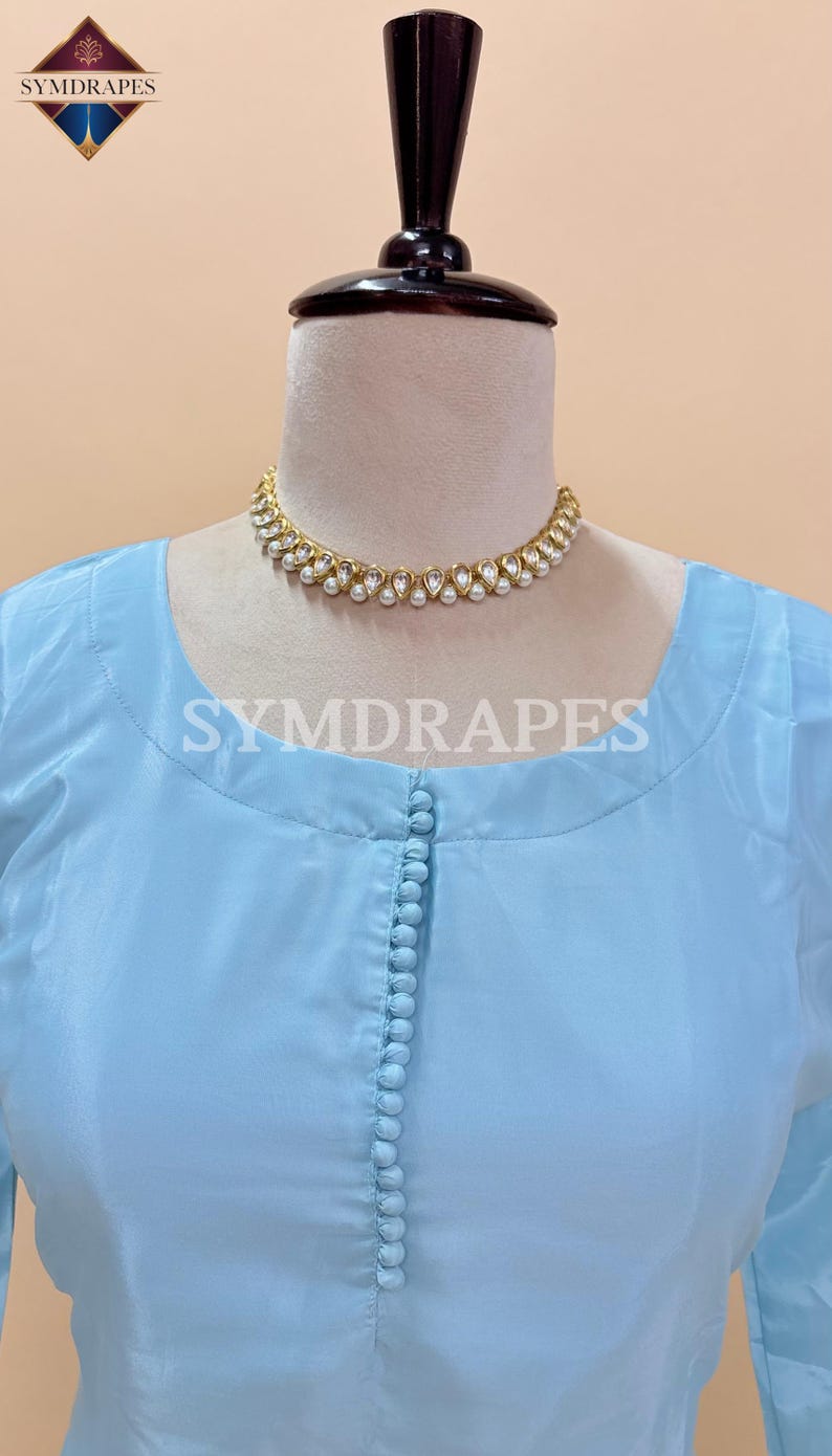 May include: A light blue tunic-style top with a row of small buttons down the front. A gold-colored necklace with white pearl accents is around the neckline. The text "SYMDRAPES" is visible on the top. The top is displayed on a mannequin.