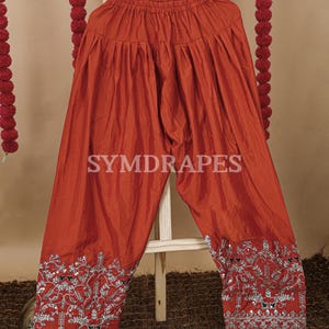 May include: Rust-colored palazzo pants with intricate silver embroidery along the hems. The pants are made of a shiny fabric and feature the text "SYMDRAPES". The pants are displayed on a wooden hanger.