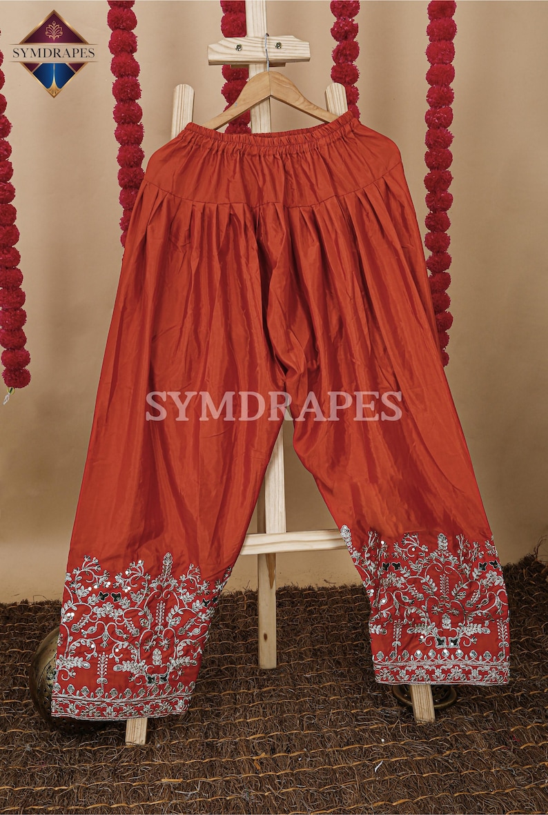 May include: Rust-colored palazzo pants with intricate silver embroidery along the hems. The pants are made of a shiny fabric and feature the text "SYMDRAPES". The pants are displayed on a wooden hanger.