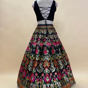 May include: A black crop top with a lace-up back paired with a long, flared skirt. The skirt features a black background with a gold floral pattern and accents of pink, yellow, and green. The hemline has a decorative border.