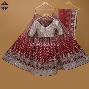 Puede incluir: Un conjunto formal con una falda rojo oscuro y un top a juego con intrincados detalles de espejo. El top tiene cuello en V y mangas largas. El conjunto incluye una dupatta a juego. La marca "SYMDRAPES" es visible.