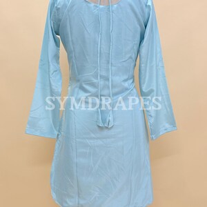 May include: A light blue tunic-style dress with long sleeves, displayed on a mannequin. The dress features a scoop neckline and a tie detail at the back. The brand name "SYMDRAPES" is printed on the garment.