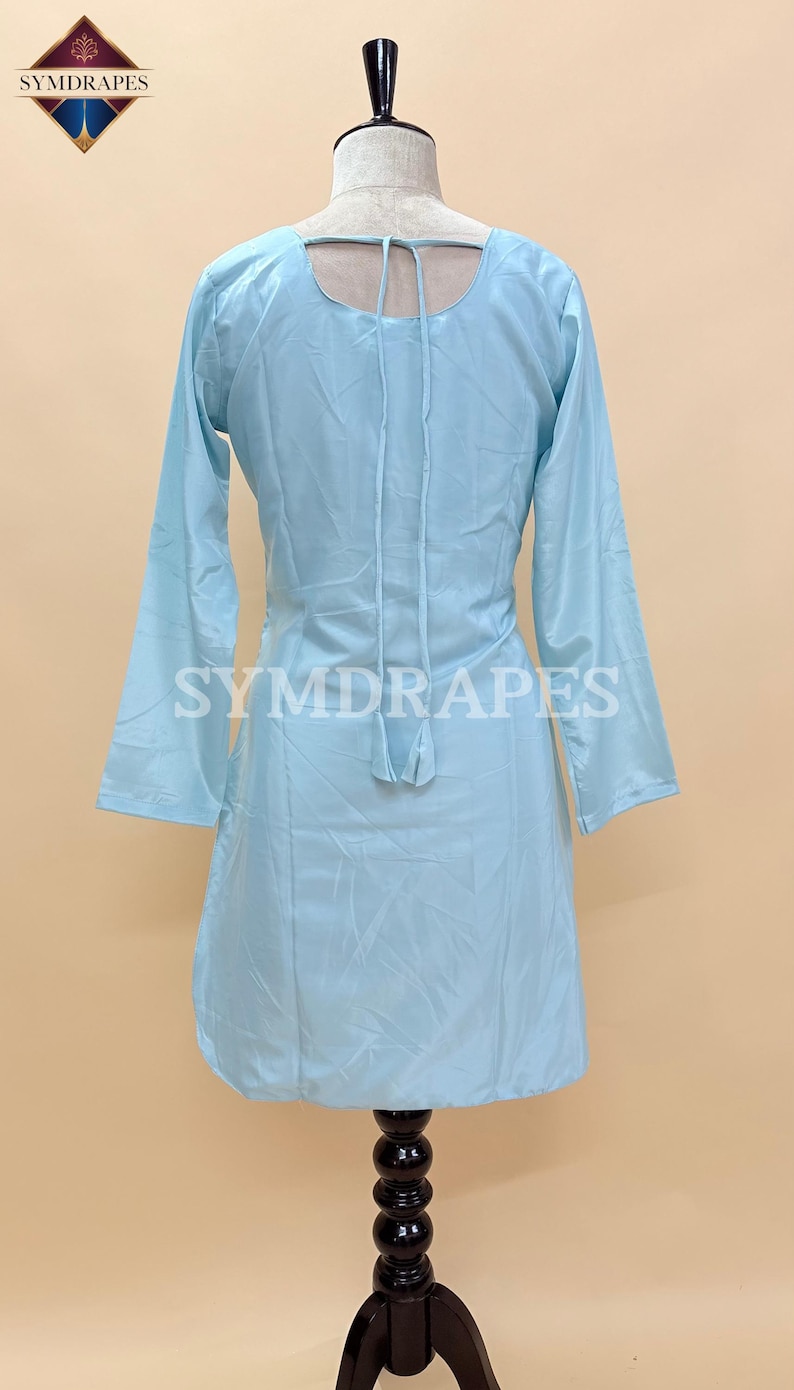 May include: A light blue tunic-style dress with long sleeves, displayed on a mannequin. The dress features a scoop neckline and a tie detail at the back. The brand name "SYMDRAPES" is printed on the garment.