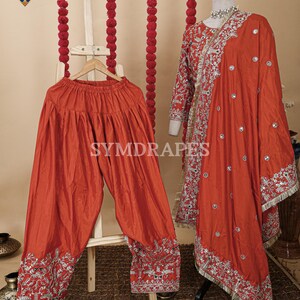 May include: A rust-colored, traditional Indian outfit, including loose-fitting pants and a long-sleeved top with a matching scarf. The outfit features intricate silver embroidery along the edges and hems. The outfit is displayed on a mannequin and a wooden hanger.