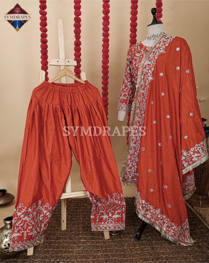 May include: A rust-colored, traditional Indian outfit, including loose-fitting pants and a long-sleeved top with a matching scarf. The outfit features intricate silver embroidery along the edges and hems. The outfit is displayed on a mannequin and a wooden hanger.