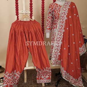 May include: A rust-colored salwar kameez set. The ensemble includes loose-fitting pants, a long-sleeved tunic, and a matching scarf. The tunic and scarf are adorned with intricate silver embroidery and mirror accents. The set is displayed on a mannequin and a wooden hanger.