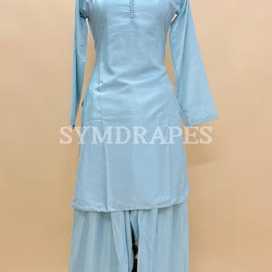 May include: A light blue, two-piece outfit, consisting of a long-sleeved tunic and wide-leg pants, displayed on a mannequin. The tunic features a round neckline and button details. The brand name "SYMDRAPES" is visible.