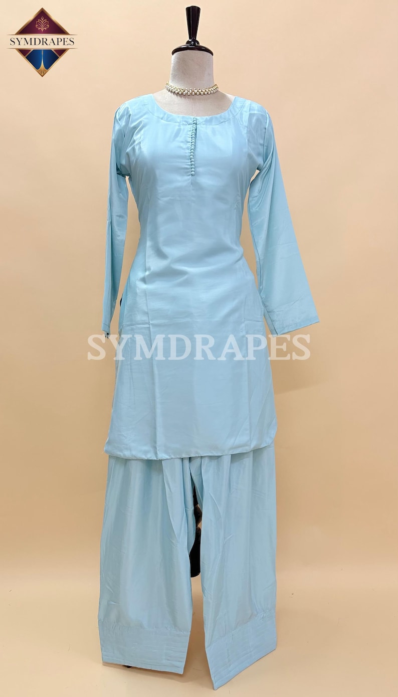 May include: A light blue, two-piece outfit, consisting of a long-sleeved tunic and wide-leg pants, displayed on a mannequin. The tunic features a round neckline and button details. The brand name "SYMDRAPES" is visible.
