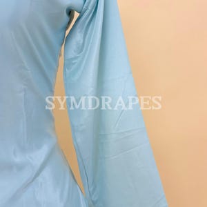 May include: Light blue long-sleeved garment with the text "SYMDRAPES" in white. The fabric has a subtle sheen, and the garment appears to be made of a lightweight material. The overall style suggests a flowing design, possibly a dress or a top.