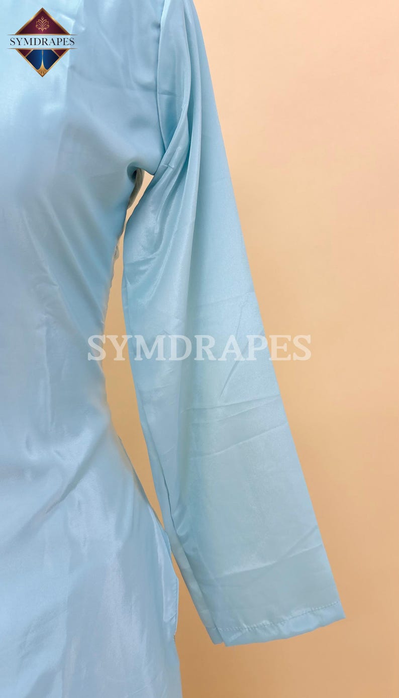 May include: Light blue long-sleeved garment with the text "SYMDRAPES" in white. The fabric has a subtle sheen, and the garment appears to be made of a lightweight material. The overall style suggests a flowing design, possibly a dress or a top.