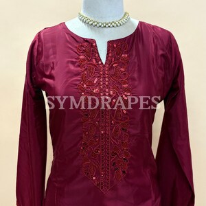 May include: A burgundy-colored tunic with long sleeves and a V-neckline. The tunic features intricate embroidery and sequin embellishments around the neckline. The brand name "SYMDRAPES" is visible on the garment.