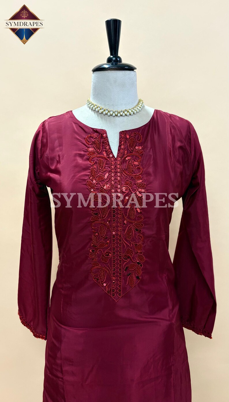May include: A burgundy-colored tunic with long sleeves and a V-neckline. The tunic features intricate embroidery and sequin embellishments around the neckline. The brand name "SYMDRAPES" is visible on the garment.