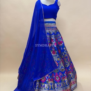Puede incluir: Un vibrante conjunto lehenga azul. El atuendo presenta un top corto sin mangas, una falda fluida con motivos florales y de elefantes, y una dupatta transparente a juego. El borde de la falda tiene un ribete metálico plateado y rosa. El logotipo "SYMDRAPES" es visible.