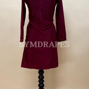 May include: A burgundy-colored long-sleeve tunic dress displayed on a black mannequin. The dress features a scoop neckline and decorative trim on the sleeves. The brand name "SYMDRAPES" is visible on the garment. The dress is made of a smooth, flowing fabric.