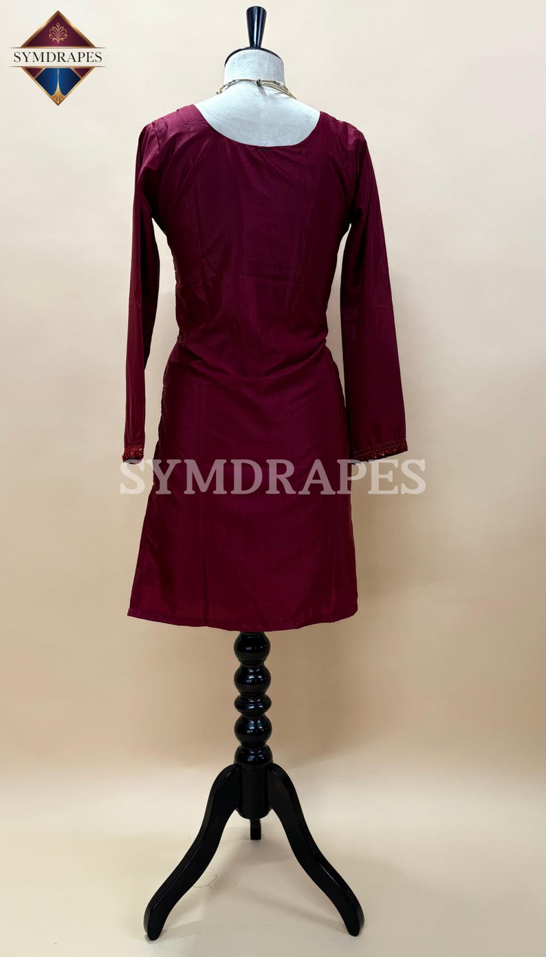 May include: A burgundy-colored long-sleeve tunic dress displayed on a black mannequin. The dress features a scoop neckline and decorative trim on the sleeves. The brand name "SYMDRAPES" is visible on the garment. The dress is made of a smooth, flowing fabric.