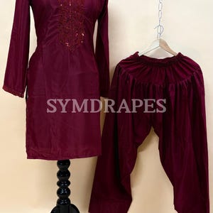 May include: A burgundy two-piece outfit, consisting of a long-sleeved tunic with embroidered detailing and matching pants. The tunic is displayed on a mannequin, and the pants are hanging on a hanger. The outfit is made of a shiny fabric. The brand name "SYMDRAPES" is visible.