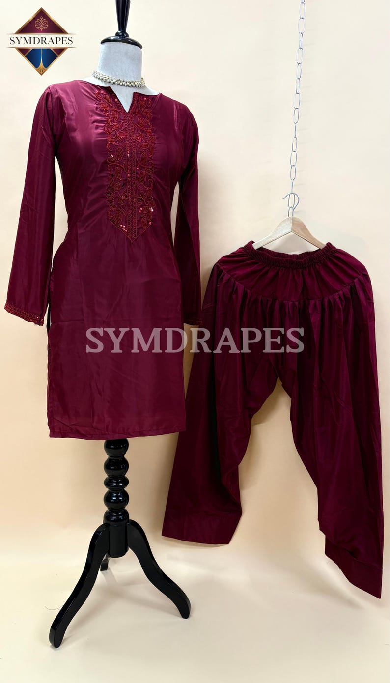 May include: A burgundy two-piece outfit, consisting of a long-sleeved tunic with embroidered detailing and matching pants. The tunic is displayed on a mannequin, and the pants are hanging on a hanger. The outfit is made of a shiny fabric. The brand name "SYMDRAPES" is visible.