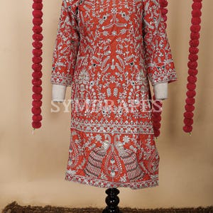 May include: A red embroidered tunic with silver embellishments and a three-quarter sleeve design. The tunic features intricate floral and peacock patterns, displayed on a black mannequin. The word "FRONT" is visible at the top.