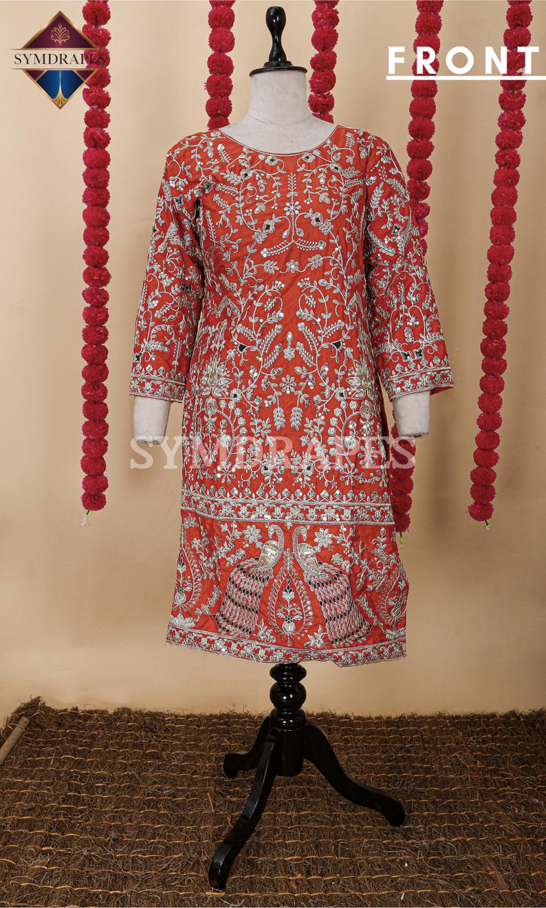 May include: A red embroidered tunic with silver embellishments and a three-quarter sleeve design. The tunic features intricate floral and peacock patterns, displayed on a black mannequin. The word "FRONT" is visible at the top.