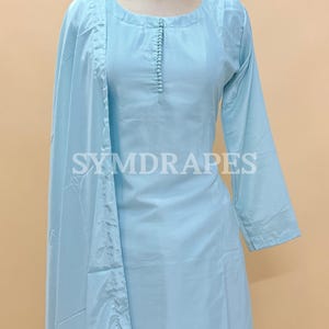 May include: A light blue, long-sleeved tunic and matching pants set. The tunic has a round neckline and a row of small buttons down the front. The set is displayed on a mannequin. The text "SYMDRAPES" is visible on the tunic and the logo is in the top left corner.