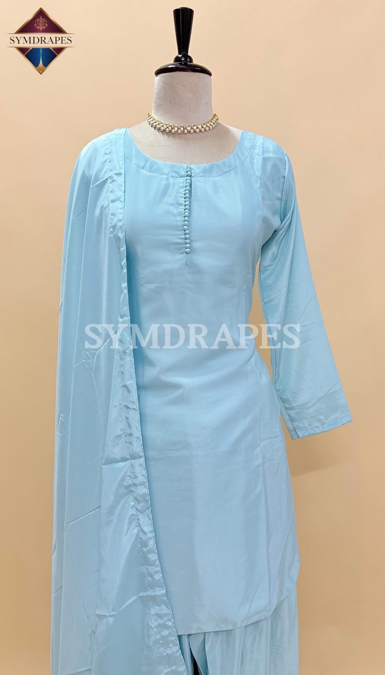 May include: A light blue, long-sleeved tunic and matching pants set. The tunic has a round neckline and a row of small buttons down the front. The set is displayed on a mannequin. The text "SYMDRAPES" is visible on the tunic and the logo is in the top left corner.