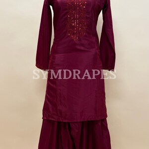 May include: A burgundy salwar kameez outfit on a mannequin. The long-sleeved tunic features intricate embroidery around the neckline and cuffs. The matching pants are wide-legged. The brand name "SYMDRAPES" is visible on the outfit.