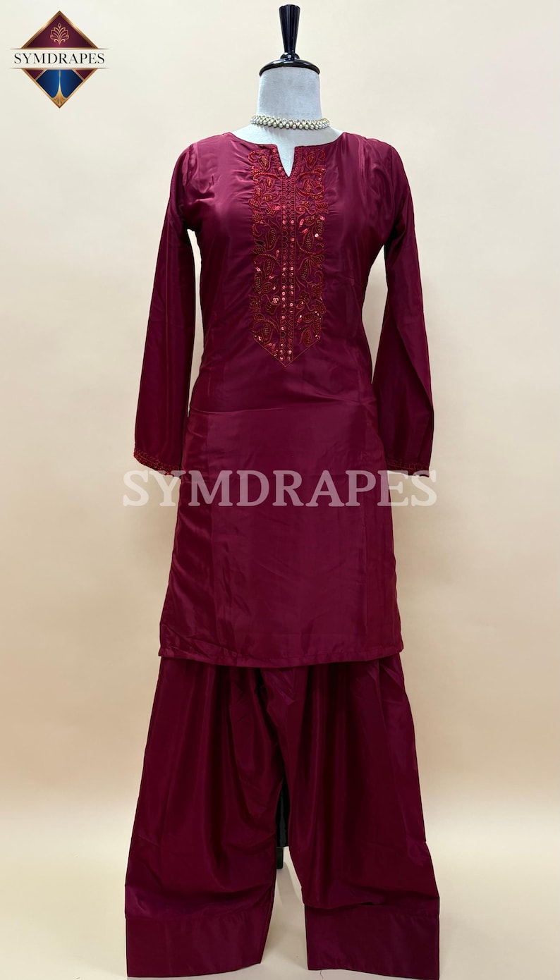 May include: A burgundy salwar kameez outfit on a mannequin. The long-sleeved tunic features intricate embroidery around the neckline and cuffs. The matching pants are wide-legged. The brand name "SYMDRAPES" is visible on the outfit.