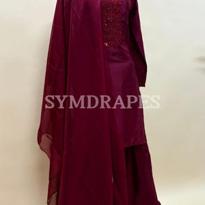 May include: A burgundy-colored, long-sleeved tunic and matching pants set with a sheer scarf. The tunic features an embroidered neckline. The brand name "SYMDRAPES" is visible on the image.