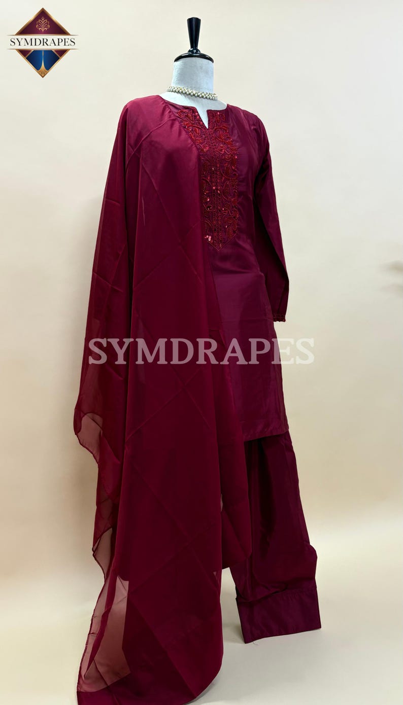May include: A burgundy-colored, long-sleeved tunic and matching pants set with a sheer scarf. The tunic features an embroidered neckline. The brand name "SYMDRAPES" is visible on the image.