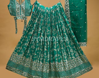 May include: A three-piece emerald green Indian outfit, including a sleeveless top, a long skirt, and a matching scarf. The skirt and top feature a floral pattern with silver accents. The outfit is displayed on wooden hangers against a neutral background.