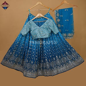 May include: A teal-colored, three-piece Indian outfit with intricate silver embroidery. The set includes a skirt, a short-sleeved top, and a matching scarf. The top has a V-neck and the skirt has a wide, flowing design. The brand name "SYMDRAPES" is visible.