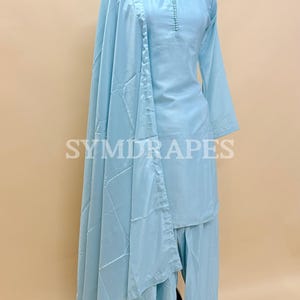 May include: A light blue, three-piece outfit on a mannequin. The set includes a long-sleeved top with button details, wide-leg pants, and a matching shawl. The outfit is displayed on a mannequin with a pearl necklace. The brand name "SYMDRAPES" is visible.
