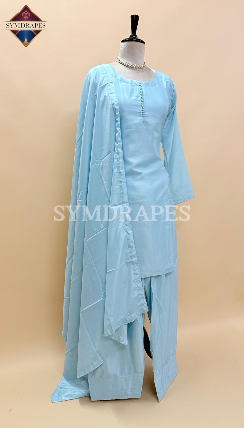May include: A light blue, three-piece outfit on a mannequin. The set includes a long-sleeved top with button details, wide-leg pants, and a matching shawl. The outfit is displayed on a mannequin with a pearl necklace. The brand name "SYMDRAPES" is visible.