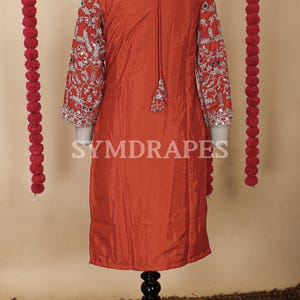 May include: A rust-colored tunic dress with intricate silver embroidery on the sleeves. The dress features a tassel detail at the neckline and is displayed on a mannequin. The background includes decorative red garlands.