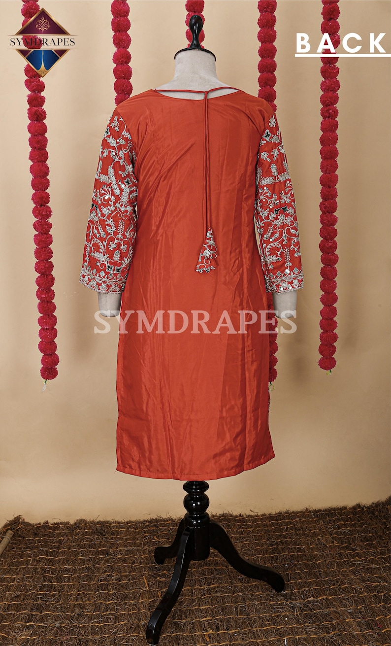May include: A rust-colored tunic dress with intricate silver embroidery on the sleeves. The dress features a tassel detail at the neckline and is displayed on a mannequin. The background includes decorative red garlands.