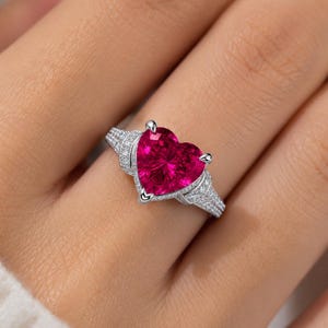 May include: A silver-toned ring featuring a large, faceted, heart-shaped vibrant pink gemstone as the centerpiece. The band is split into two sections on each side, encrusted with small, clear, sparkling stones that extend along the shank.