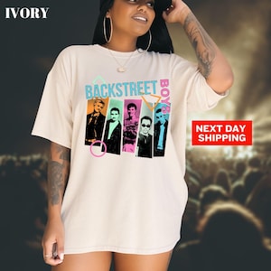 May include: Ivory-colored t-shirt featuring the text "BACKSTREET BOYS" in teal and pink. The shirt displays images of the band members in colorful vertical panels. The words "NEXT DAY SHIPPING" are in a red box.