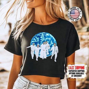 May include: Black crop top featuring a graphic of a group of people in white outfits inside a blue sphere with a geometric design. The top has a round neckline and short sleeves. A circular "Made in the USA" logo is visible.
