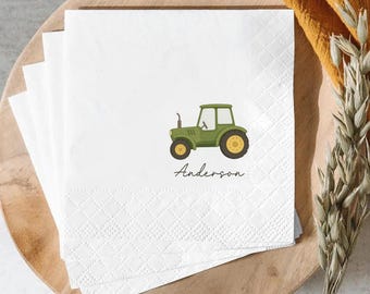 May include: A stack of white square paper napkins featuring a green tractor illustration and the name Anderson printed in black script lettering. The napkins have a textured embossed border pattern along the edges of the square paper surface.